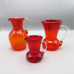 May include: Set of three red glass pitchers with textured surfaces. The largest pitcher is orange and yellow, the medium pitcher is red, and the smallest pitcher is red.