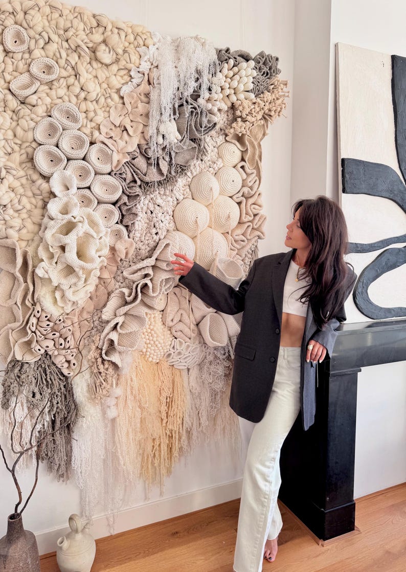 May include: A large, textured wall hanging with a variety of neutral-toned, handcrafted elements. The artwork features a mix of woven, crocheted, and knotted textures in shades of cream, beige, and gray. The piece is displayed on a white wall.