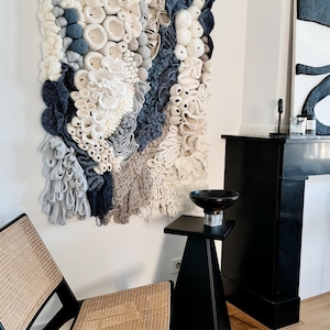 Puede incluir: Un gran tapiz de pared texturizado con una variedad de elementos de ganchillo y tejido en blanco roto, gris y azul marino. La obra de arte se exhibe en una pared blanca, con una mesa auxiliar negra y una silla con asiento tejido en primer plano.