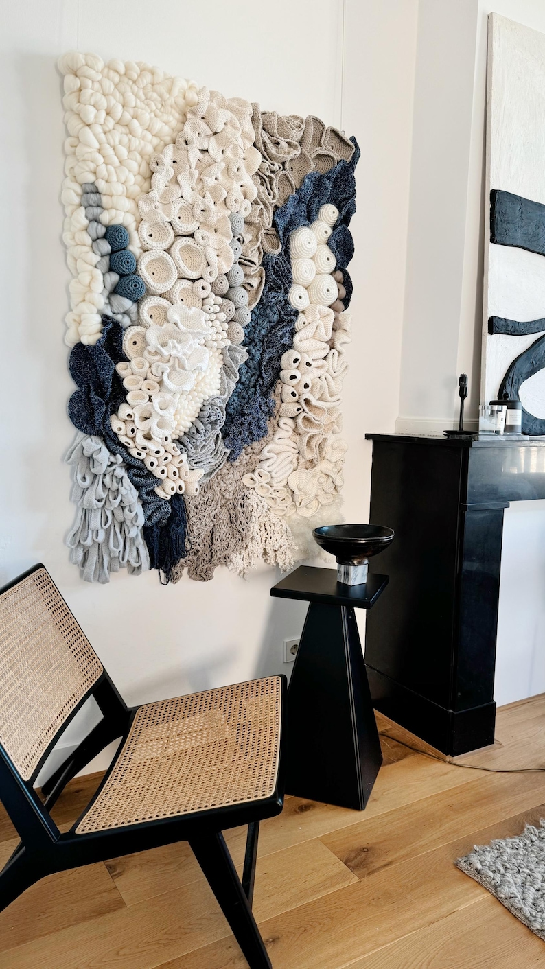 Puede incluir: Un gran tapiz de pared texturizado con una variedad de elementos de ganchillo y tejido en blanco roto, gris y azul marino. La obra de arte se exhibe en una pared blanca, con una mesa auxiliar negra y una silla con asiento tejido en primer plano.