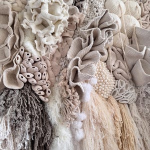 May include: A textured wall hanging featuring a variety of neutral-toned, handcrafted elements. The artwork incorporates various textures, including crochet, woven, and fringed details, creating a tactile and visually rich composition. The color palette includes shades of beige, cream, and gray.