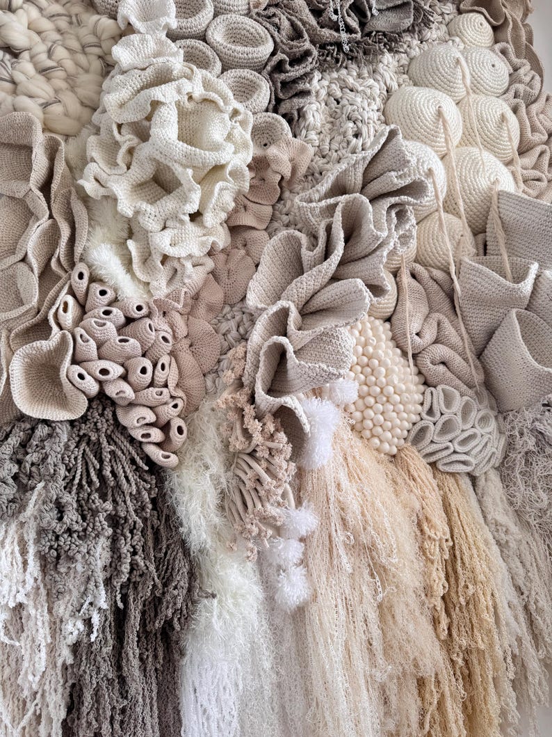 May include: A textured wall hanging featuring a variety of neutral-toned, handcrafted elements. The artwork incorporates various textures, including crochet, woven, and fringed details, creating a tactile and visually rich composition. The color palette includes shades of beige, cream, and gray.