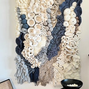Puede incluir: Un gran tapiz de pared texturizado con una variedad de elementos en blanco roto, beige y azul marino. La obra de arte presenta una mezcla de texturas tejidas y de ganchillo, creando un efecto tridimensional. La pieza est&aacute; montada en una pared blanca.