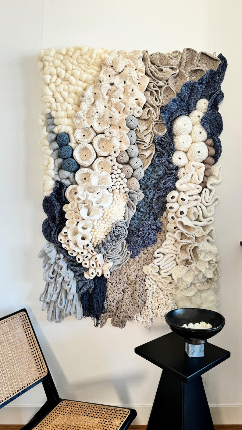 Puede incluir: Un gran tapiz de pared texturizado con una variedad de elementos en blanco roto, beige y azul marino. La obra de arte presenta una mezcla de texturas tejidas y de ganchillo, creando un efecto tridimensional. La pieza est&aacute; montada en una pared blanca.