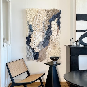 Puede incluir: Un gran tapiz de pared texturizado con una variedad de colores neutros, incluyendo blanco, beige y azul marino. La obra de arte presenta una mezcla de formas y texturas, creando una pieza &uacute;nica y art&iacute;stica. La habitaci&oacute;n tambi&eacute;n incluye una silla negra y una mesa auxiliar.