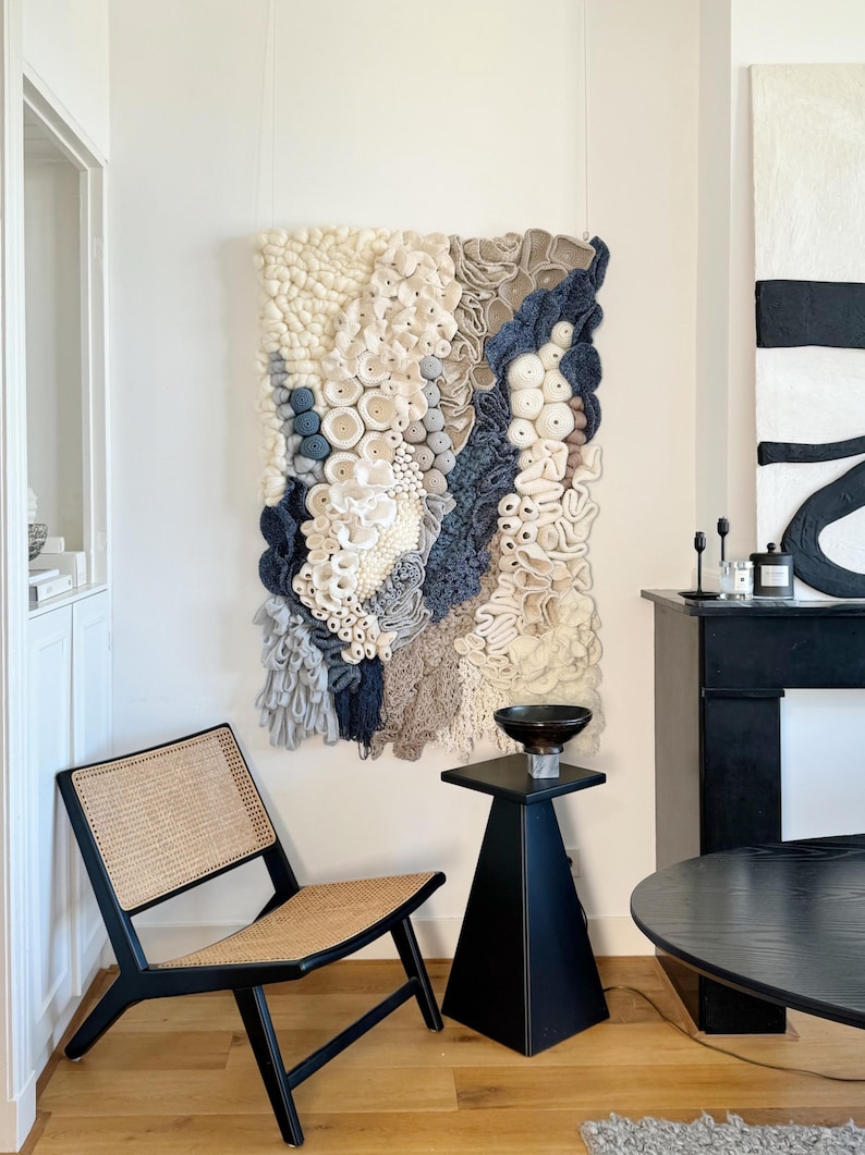 Puede incluir: Un gran tapiz de pared texturizado con una variedad de colores neutros, incluyendo blanco, beige y azul marino. La obra de arte presenta una mezcla de formas y texturas, creando una pieza &uacute;nica y art&iacute;stica. La habitaci&oacute;n tambi&eacute;n incluye una silla negra y una mesa auxiliar.