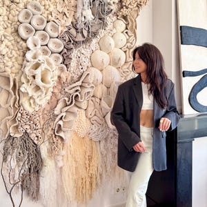 May include: A large, textured wall hanging with a variety of neutral tones and shapes. The artwork features a mix of woven, knotted, and fringed elements, creating a tactile and organic aesthetic. The piece is mounted on a white wall.