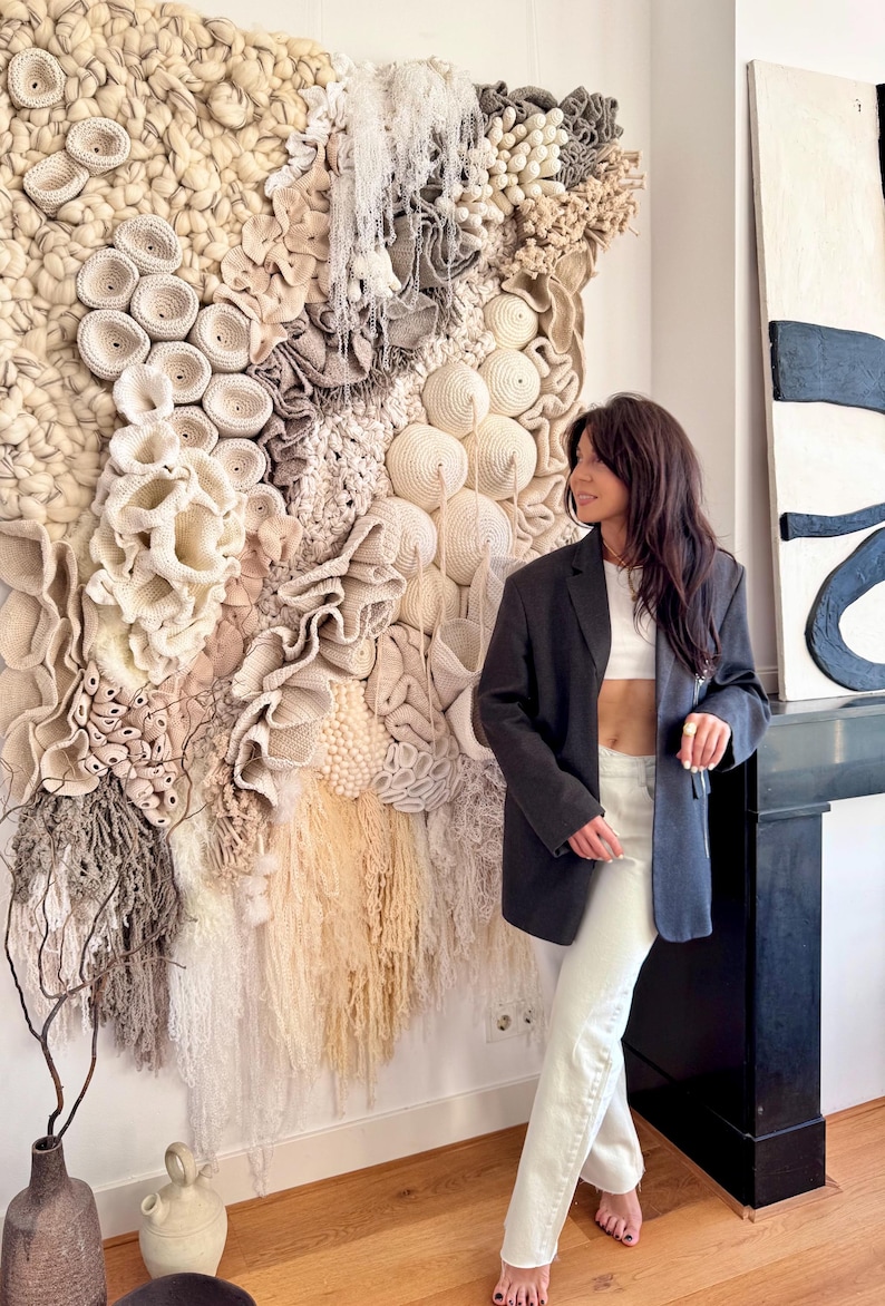 May include: A large, textured wall hanging with a variety of neutral tones and shapes. The artwork features a mix of woven, knotted, and fringed elements, creating a tactile and organic aesthetic. The piece is mounted on a white wall.