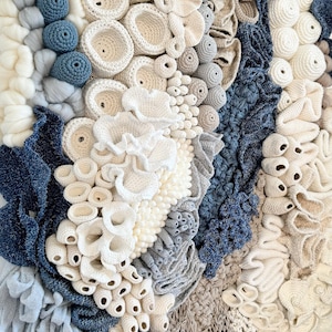 Puede incluir: Obra de arte textil con diversas texturas y colores. La obra presenta una combinaci&oacute;n de elementos de ganchillo, tejido y anudado en tonos blancos, crema, grises y azules. El dise&ntilde;o evoca formas y texturas org&aacute;nicas.