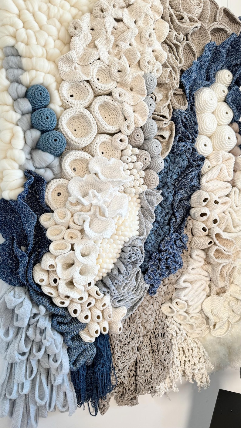 Puede incluir: Obra de arte textil con diversas texturas y colores. La obra presenta una combinaci&oacute;n de elementos de ganchillo, tejido y anudado en tonos blancos, crema, grises y azules. El dise&ntilde;o evoca formas y texturas org&aacute;nicas.