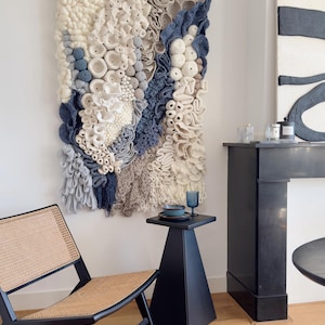 Puede incluir: Un gran tapiz de pared texturizado con un dise&ntilde;o de arrecife de coral en tonos blancos, azules y grises. La obra de arte se exhibe en un espacio de vida moderno con una mesa auxiliar, una silla y una chimenea negras. La pieza tejida a&ntilde;ade un toque art&iacute;stico &uacute;nico.