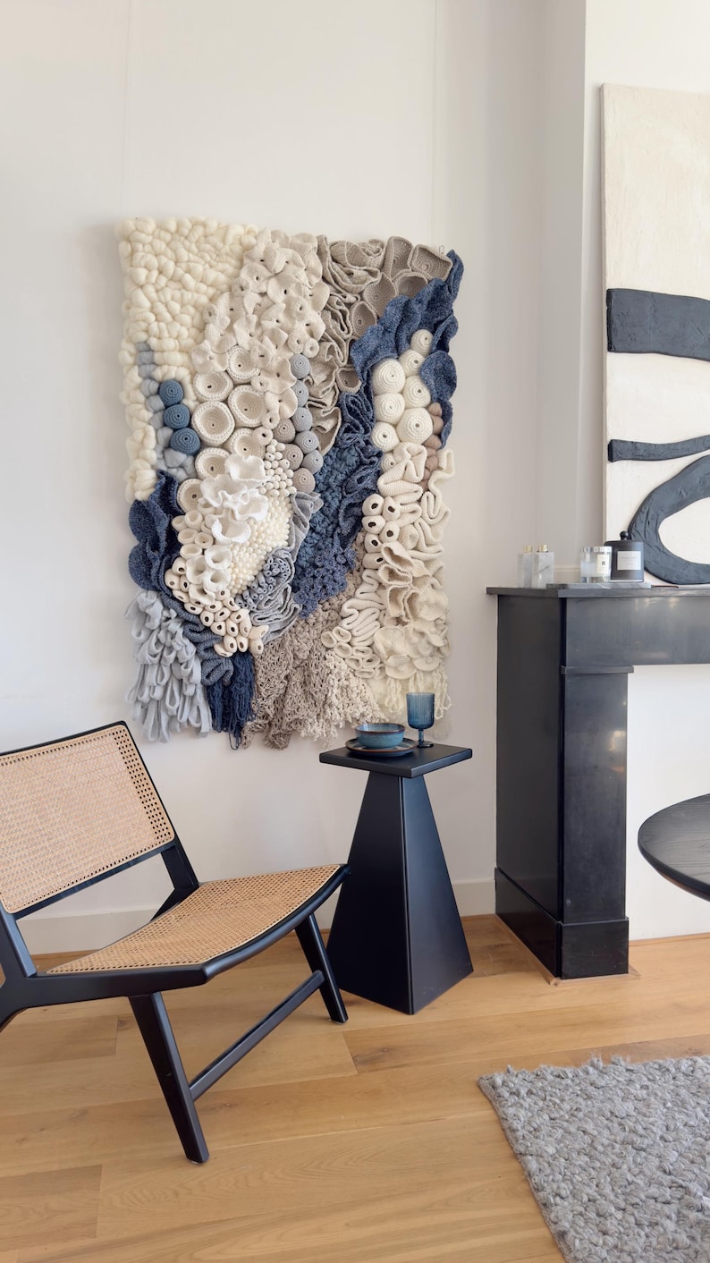 Puede incluir: Un gran tapiz de pared texturizado con un dise&ntilde;o de arrecife de coral en tonos blancos, azules y grises. La obra de arte se exhibe en un espacio de vida moderno con una mesa auxiliar, una silla y una chimenea negras. La pieza tejida a&ntilde;ade un toque art&iacute;stico &uacute;nico.