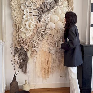 May include: A large, textured wall hanging with a variety of neutral-toned, handcrafted elements. The artwork features circular, ruffled, and fringed details in shades of cream, beige, and gray. The piece is displayed on a white wall, creating a focal point in the room.