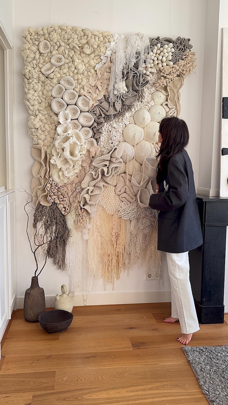 May include: A large, textured wall hanging with a variety of neutral-toned, handcrafted elements. The artwork features circular, ruffled, and fringed details in shades of cream, beige, and gray. The piece is displayed on a white wall, creating a focal point in the room.