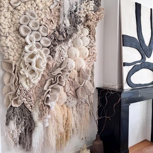 May include: A large, textured wall hanging with a variety of neutral tones, including beige, cream, and gray. The artwork features a mix of woven, crocheted, and fringed elements, creating a 3D effect. The piece is mounted on a white wall.