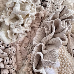 May include: Close-up of a textured textile art piece. The artwork features various crochet and knitted elements in shades of cream, beige, and light brown. The textures include ruffled edges, round shapes, and tufted details, creating a tactile and layered composition.