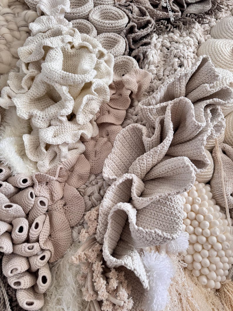 May include: Close-up of a textured textile art piece. The artwork features various crochet and knitted elements in shades of cream, beige, and light brown. The textures include ruffled edges, round shapes, and tufted details, creating a tactile and layered composition.