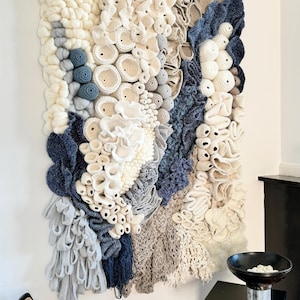 Puede incluir: Tapiz de pared texturizado con una variedad de elementos tejidos y de ganchillo en blanco roto, beige y azul marino. La obra de arte tiene una apariencia tridimensional y org&aacute;nica, que se asemeja a corales o formas abstractas. Montado en una pared blanca.