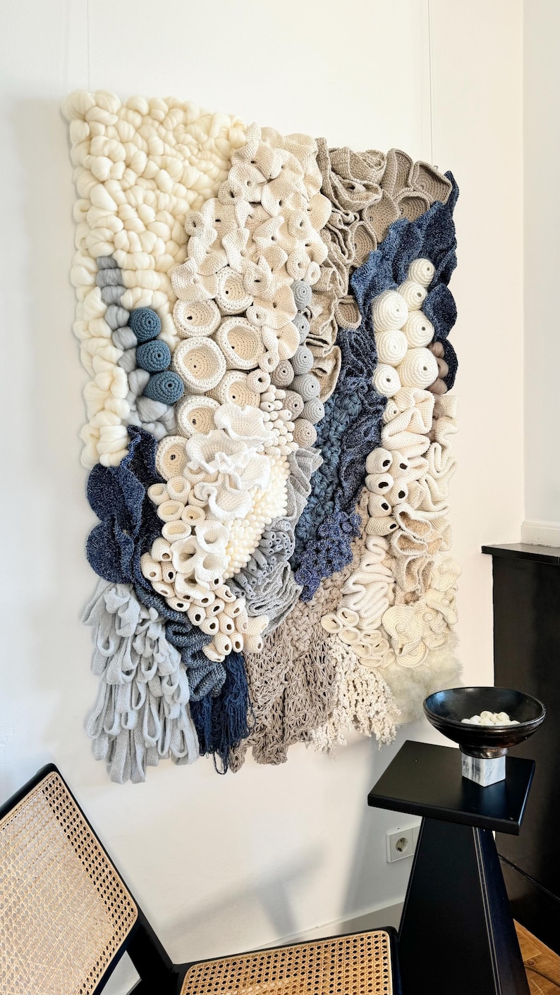 Puede incluir: Tapiz de pared texturizado con una variedad de elementos tejidos y de ganchillo en blanco roto, beige y azul marino. La obra de arte tiene una apariencia tridimensional y org&aacute;nica, que se asemeja a corales o formas abstractas. Montado en una pared blanca.