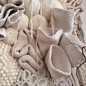 May include: A textured, abstract wall hanging composed of various off-white and beige crocheted elements. The piece features spherical shapes, basket-like forms, and intricate, layered textures, creating a tactile and visually engaging artwork. The neutral color palette adds to its organic aesthetic.