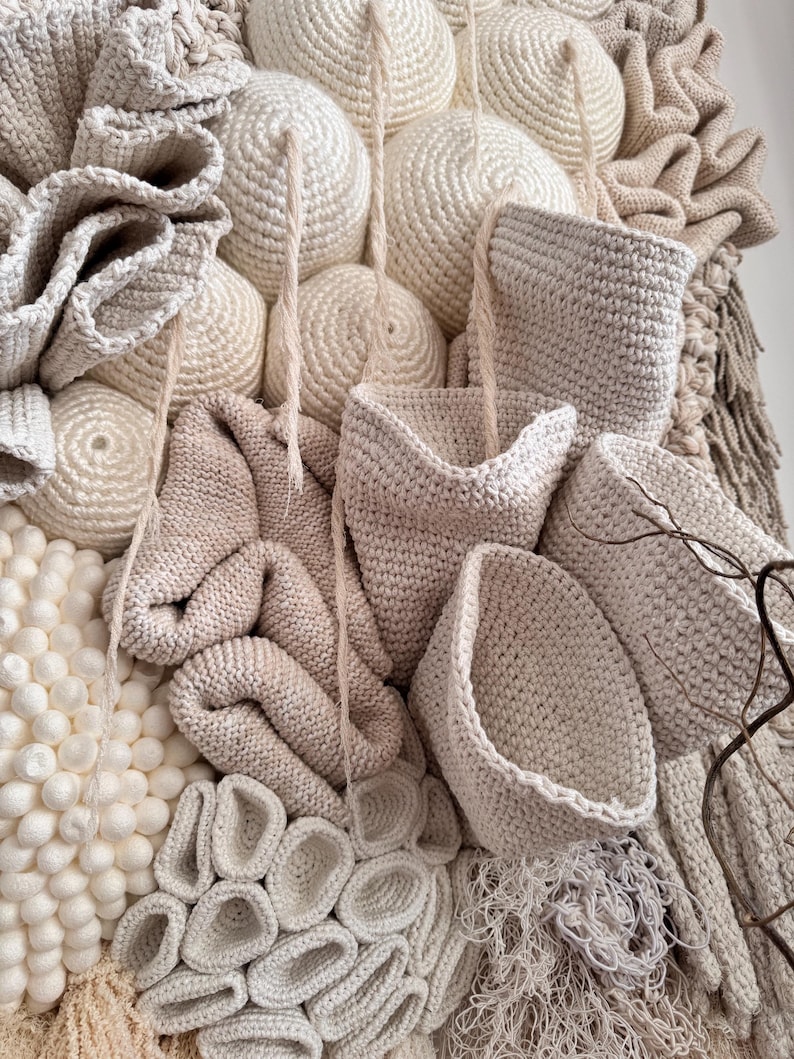 May include: A textured, abstract wall hanging composed of various off-white and beige crocheted elements. The piece features spherical shapes, basket-like forms, and intricate, layered textures, creating a tactile and visually engaging artwork. The neutral color palette adds to its organic aesthetic.