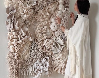 Könnte beinhalten: Ein großer, quadratischer Wandbehang mit einer komplexen, dreidimensionalen Textilkunstkomposition in Creme-, Beige- und Off-White-Tönen. Das Stück integriert verschiedene Texturen, darunter gehäkelte korallenartige Strukturen, gefilzte Wollkugeln, geknotete Fransen und gewebte Elemente, die eine dichte, organische und geschichtete Reliefoberfläche bilden.