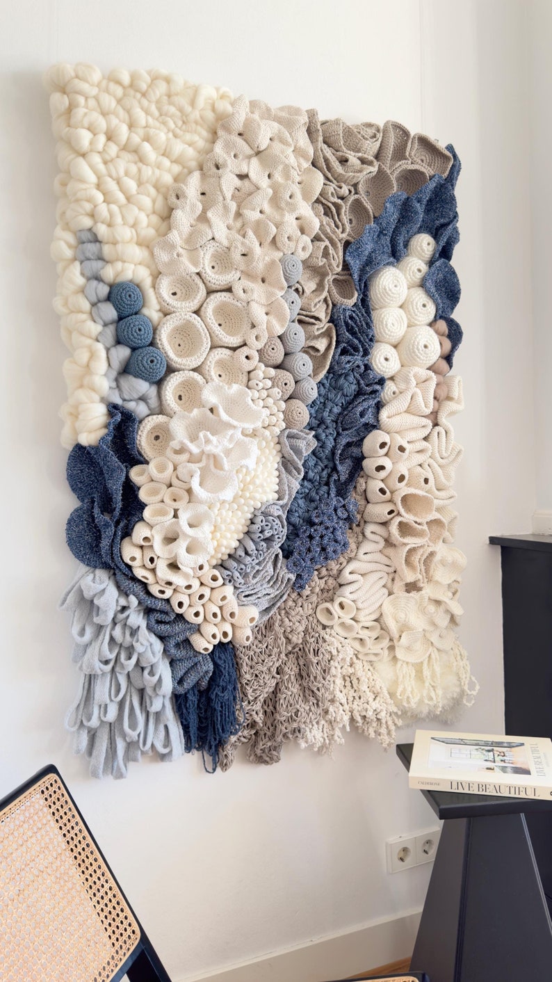 Puede incluir: Tapiz de pared texturizado con una variedad de elementos de ganchillo y tejido en blanco roto, beige y azul. La obra de arte presenta una mezcla de formas y texturas org&aacute;nicas, creando un efecto tridimensional &uacute;nico. La pieza est&aacute; montada en una pared blanca.