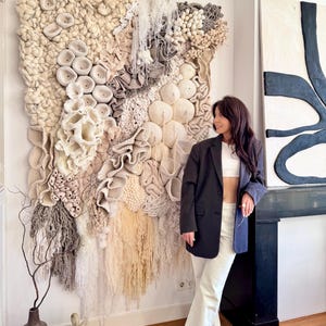 May include: A large, textured wall hanging with a variety of neutral tones, including cream, beige, and gray. The artwork features a mix of woven and sculpted elements, creating a three-dimensional effect. The piece is mounted on a white wall.