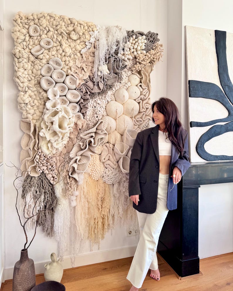 May include: A large, textured wall hanging with a variety of neutral tones, including cream, beige, and gray. The artwork features a mix of woven and sculpted elements, creating a three-dimensional effect. The piece is mounted on a white wall.