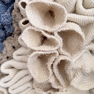 Puede incluir: Primer plano de una obra de arte textil texturizada, de color blanquecino. La obra presenta varias formas, incluyendo formas redondeadas con aberturas y estructuras en forma de p&eacute;talos superpuestas. Se ven tonos de azul y gris.