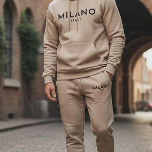 May include: A beige hoodie and matching sweatpants set. The hoodie has a drawstring hood, a front pocket, and the text "MILANO ITALY" printed on the chest. The sweatpants also have the text "MILANO ITALY" printed on the leg. The person is wearing black sneakers.