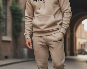 May include: A beige hoodie and matching sweatpants set. The hoodie has a drawstring hood, a front pocket, and the text "MILANO ITALY" printed on the chest. The sweatpants also have the text "MILANO ITALY" printed on the leg. The person is wearing black sneakers.