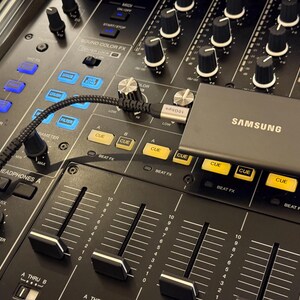 May include: A grey Samsung portable solid state drive connected via a black braided USB-C cable to a black professional DJ mixer. The mixer features numerous black rotary knobs, yellow illuminated cue buttons, and vertical faders on a dark surface.