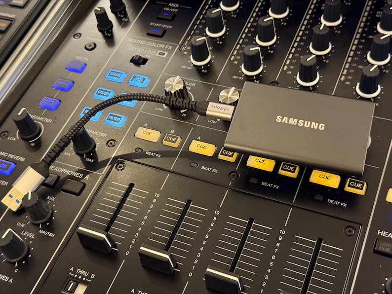 May include: A grey Samsung portable solid state drive connected via a black braided USB-C cable to a black professional DJ mixer. The mixer features numerous black rotary knobs, yellow illuminated cue buttons, and vertical faders on a dark surface.