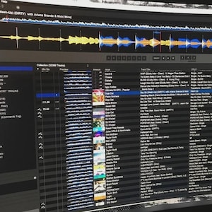 May include: A computer monitor displaying DJ software with a waveform visualization at the top and a detailed music library list below. The interface shows columns for track title, artist, album, time, and genre, with a highlighted track selection.