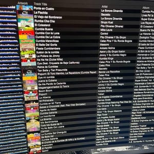 May include: A computer screen displays a music library with a list of songs, artists, and albums. The interface shows album art and track titles, with a focus on Latin music genres like Cumbia. The screen is angled.