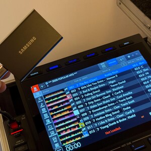 May include: A dark gray Samsung portable solid state drive connected via a silver USB-C cable to a black DJ controller. The controller features a bright digital display screen showing a music playlist with track titles, artist names, and waveforms.
