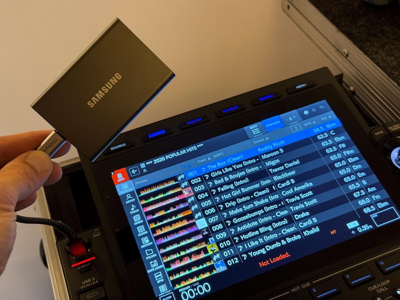 May include: A dark gray Samsung portable solid state drive connected via a silver USB-C cable to a black DJ controller. The controller features a bright digital display screen showing a music playlist with track titles, artist names, and waveforms.