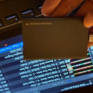 May include: A dark gray Samsung Portable SSD T7 external drive with a silver and black USB-C connector attached to the side. The device is rectangular with rounded edges and features the brand name and model printed in white text on the surface.
