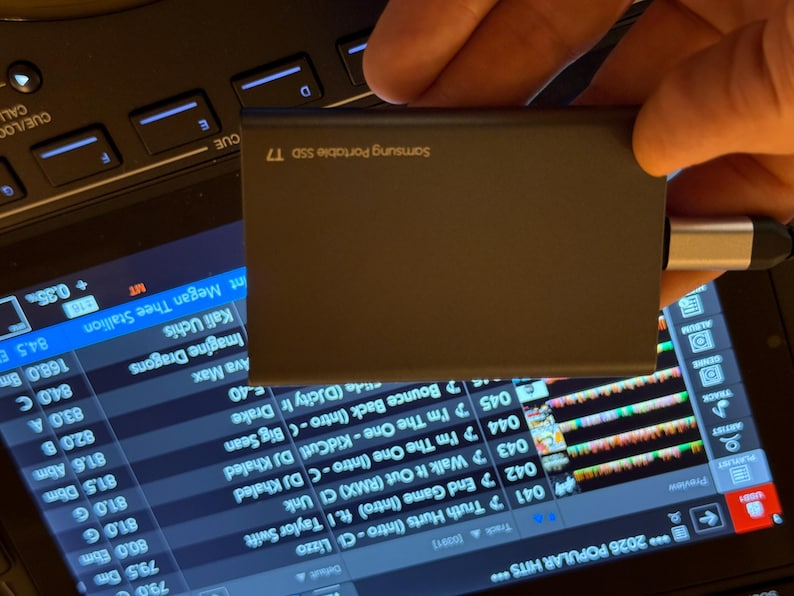 May include: A dark gray Samsung Portable SSD T7 external drive with a silver and black USB-C connector attached to the side. The device is rectangular with rounded edges and features the brand name and model printed in white text on the surface.