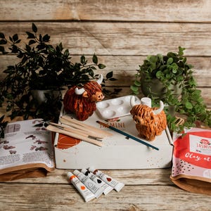 May include: A craft kit with Highland cow figurines, paint, brushes, and a palette. The kit includes a box with the word "Jasmine" and the phrase "Clay at home" on the packaging. The scene is set on a wooden surface with greenery.