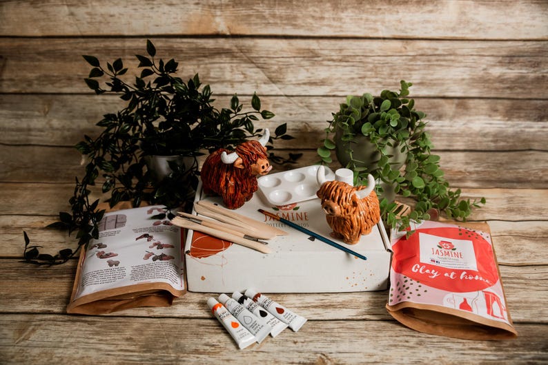May include: A craft kit with Highland cow figurines, paint, brushes, and a palette. The kit includes a box with the word "Jasmine" and the phrase "Clay at home" on the packaging. The scene is set on a wooden surface with greenery.