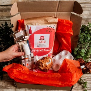 May include: A craft kit in an open cardboard box, filled with red tissue paper and various art supplies. The kit includes paint bottles, a paint palette, and a bag labeled "Jasmine Clay at Home". Decorative Highland cow figurines are also included.