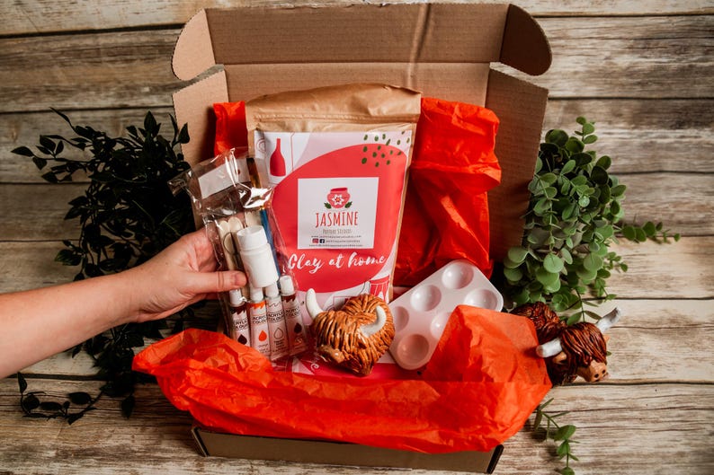 May include: A craft kit in an open cardboard box, filled with red tissue paper and various art supplies. The kit includes paint bottles, a paint palette, and a bag labeled "Jasmine Clay at Home". Decorative Highland cow figurines are also included.