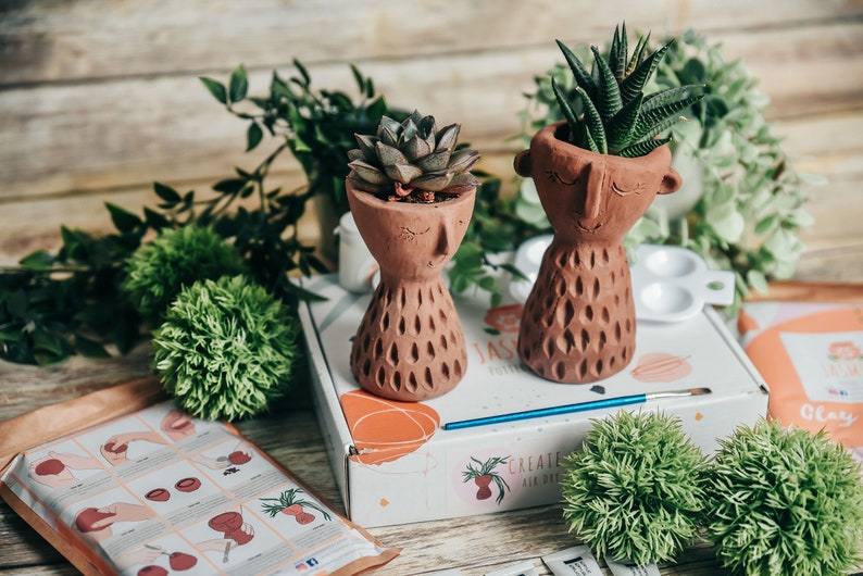 May include: Two terracotta pots shaped like faces with green succulents planted inside. The pots are sitting on a white box with the words "Create Your Own" printed on it. There are also green decorative balls and a paintbrush on the table.
