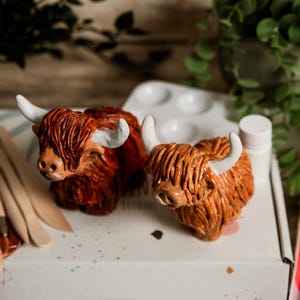 May include: Two ceramic Highland cow figurines with shaggy, reddish-brown fur and white horns. The cows are positioned on a white surface, alongside a paint palette, paint tubes, and wooden tools. The background includes greenery and a wooden surface.