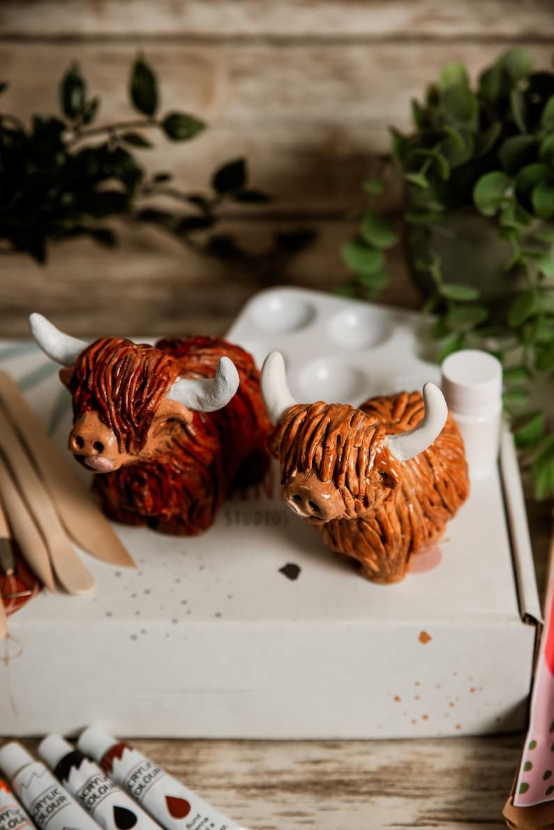 May include: Two ceramic Highland cow figurines with shaggy, reddish-brown fur and white horns. The cows are positioned on a white surface, alongside a paint palette, paint tubes, and wooden tools. The background includes greenery and a wooden surface.