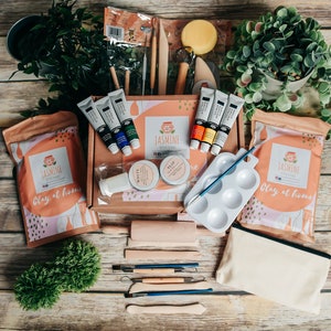 May include: A craft kit with clay, paints, and tools, arranged on a wooden surface. The kit includes paint tubes, clay bags, a paint palette, and various sculpting tools. The kit is branded with the name "Jasmine" and the phrase "Clay at home".