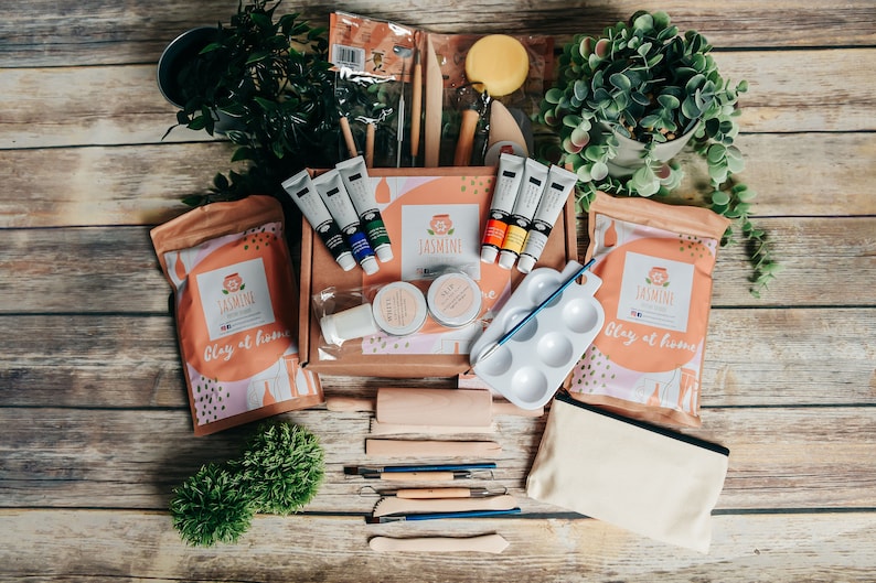 May include: A craft kit with clay, paints, and tools, arranged on a wooden surface. The kit includes paint tubes, clay bags, a paint palette, and various sculpting tools. The kit is branded with the name "Jasmine" and the phrase "Clay at home".
