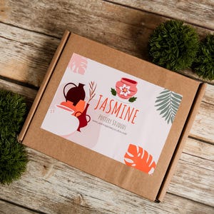 May include: A brown cardboard box with a sticker featuring pottery illustrations and the text "Jasmine Pottery Studios." The box is on a wooden surface, with two green decorative balls. The sticker has a pink background with orange and green accents.